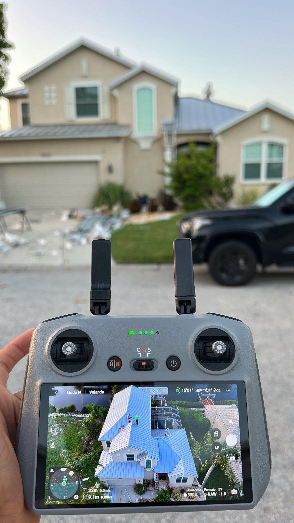 Hands holding a drone controller, viewing a suburban house on the screen and in the background.