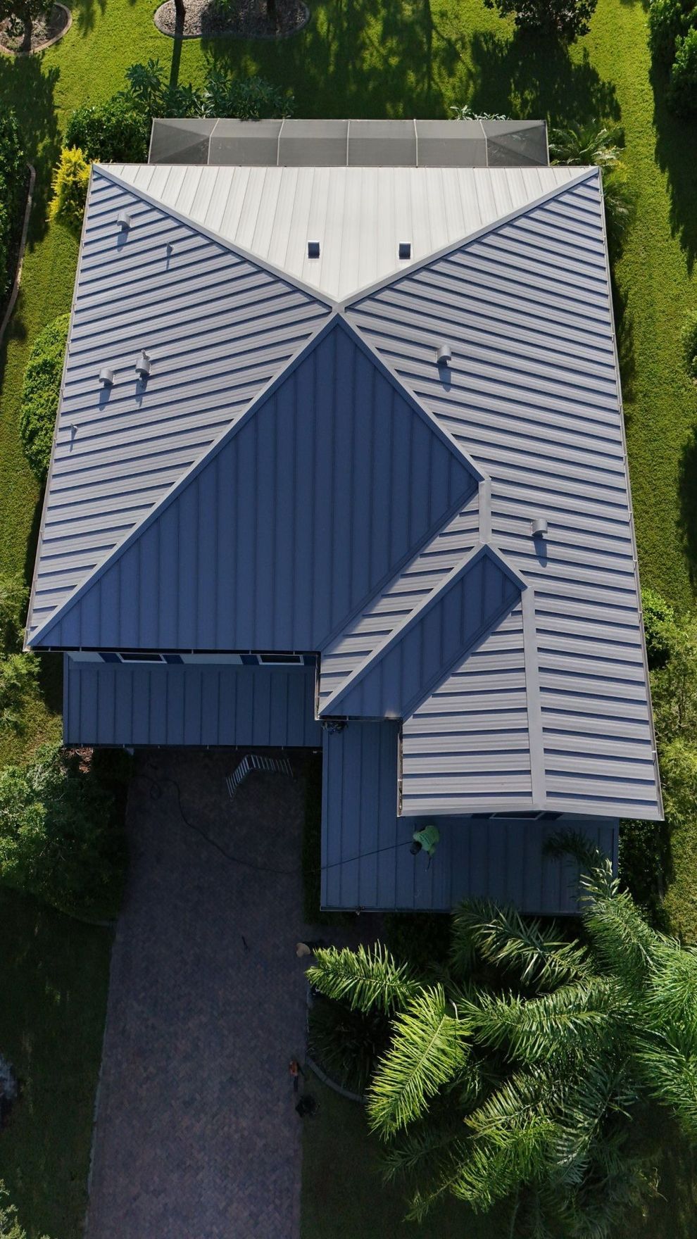 Aerial view of a house roof with gray shingles and dark blue sections, surrounded by trees and greenery.
