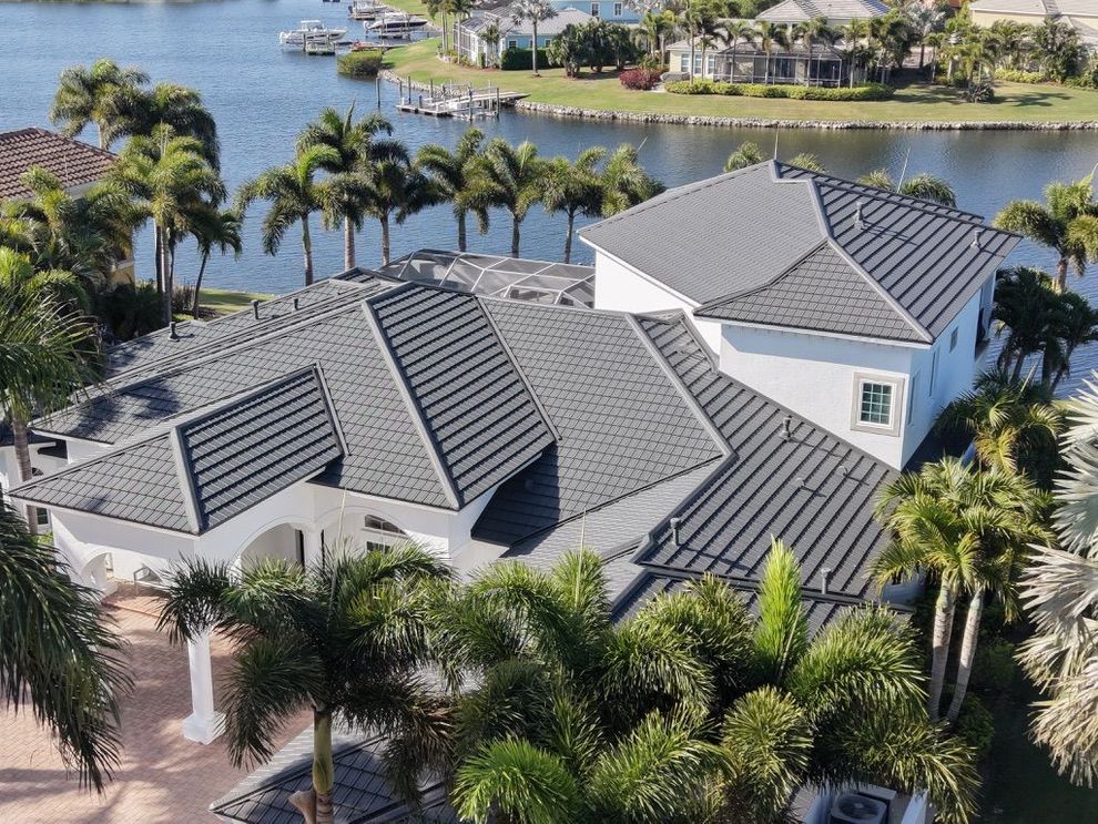 Aerial view of a gray-roofed waterfront house surrounded by palm trees and water.