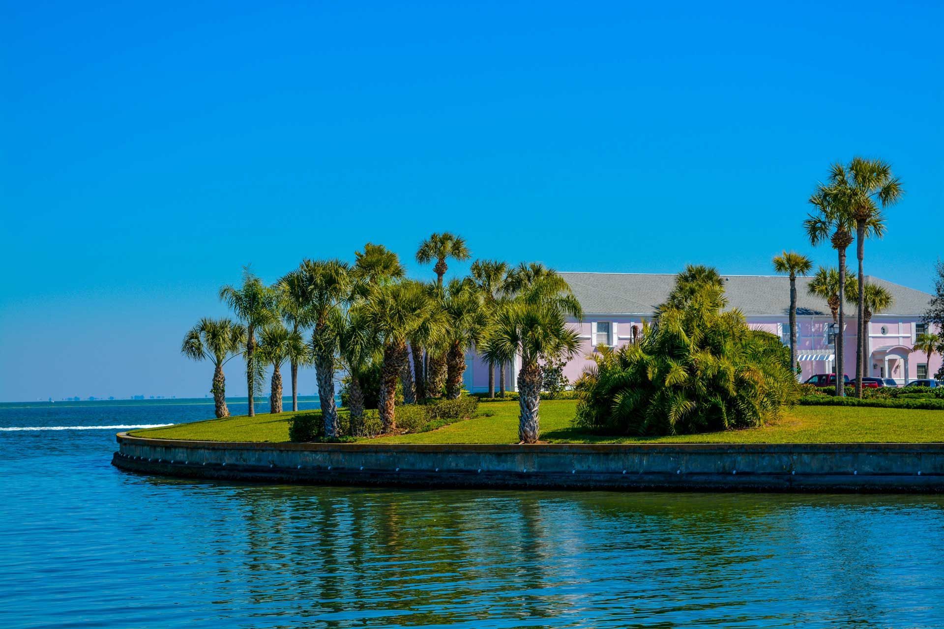Palm trees and manicured lawn by the water in front of a light-colored building on a sunny day.