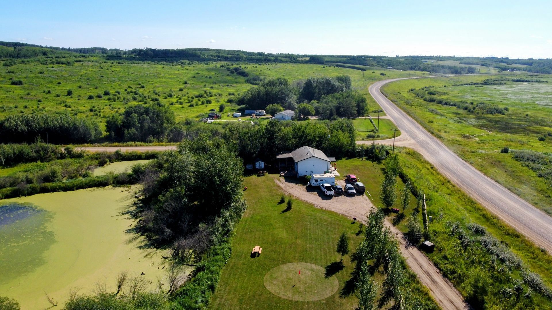 Aerial view of a white house with vehicles parked on a grassy lot next to a road, pond, and green fields.