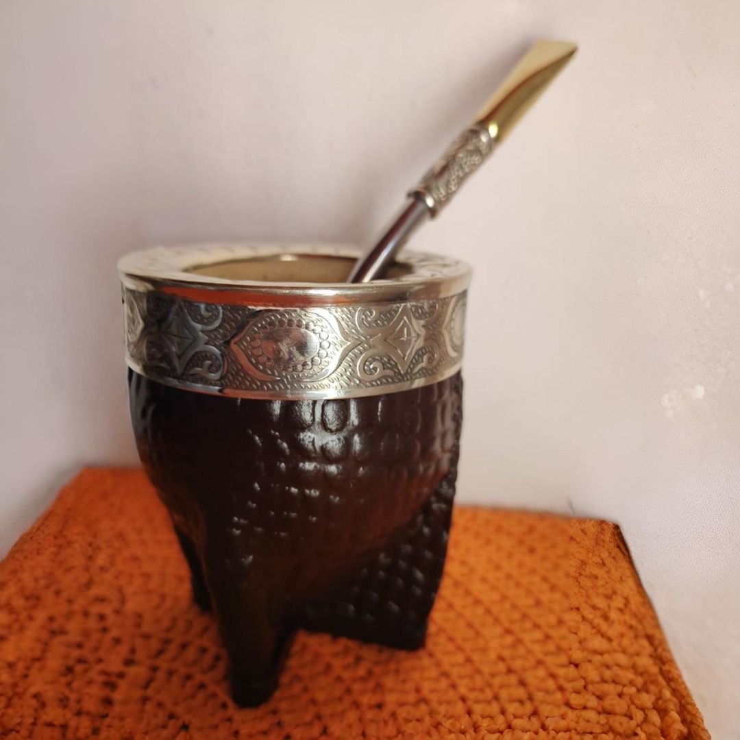 Mate de color marrón oscuro con borde plateado y pajita, colocado sobre una estera naranja.