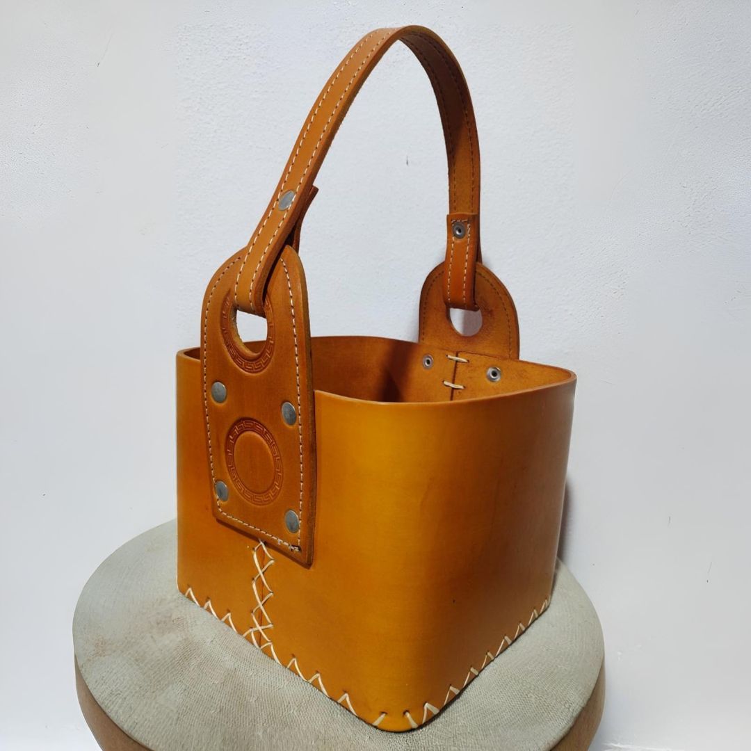 Bolso de mano de piel color tostado con asa curva, remaches y costuras blancas.