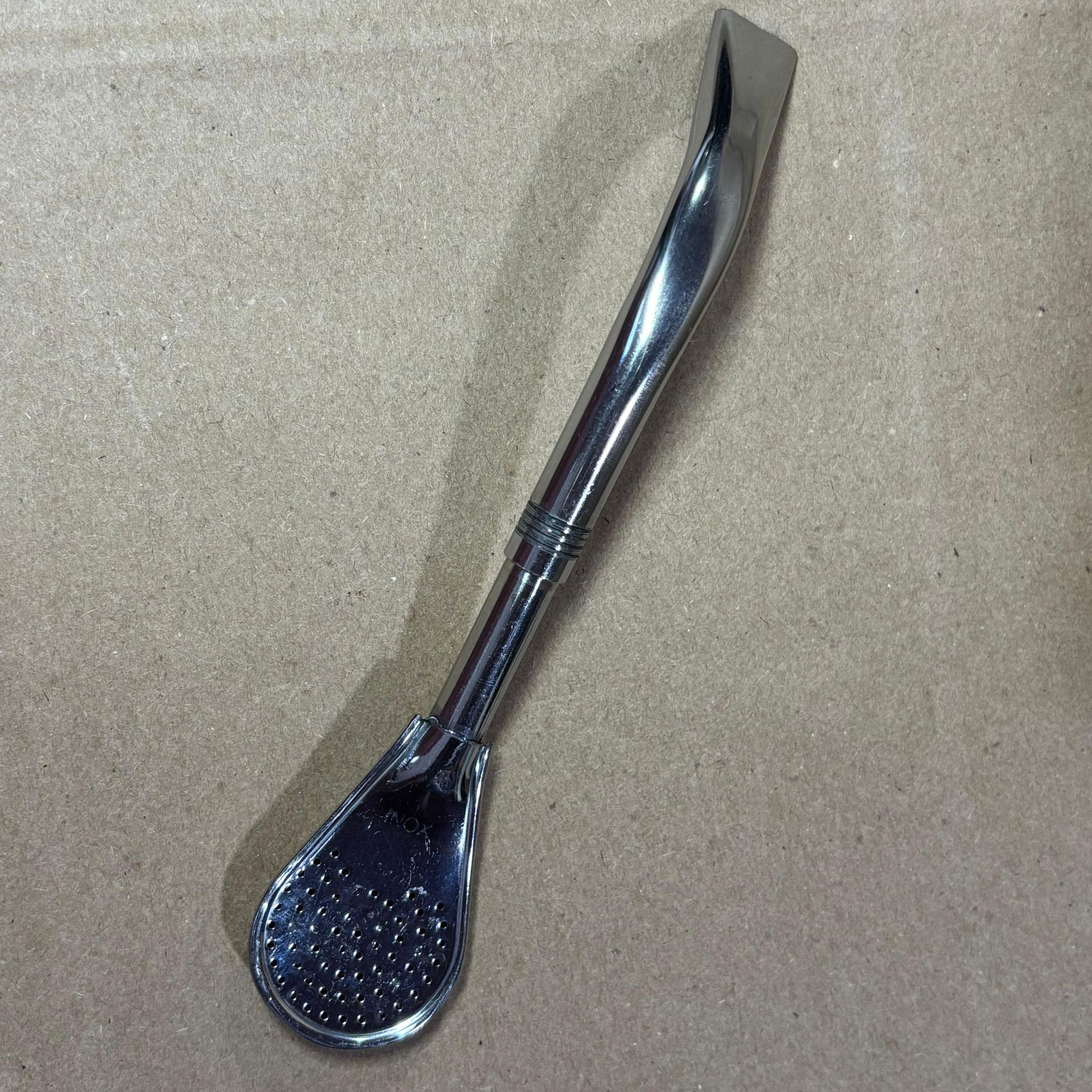 Bombilla de plata con extremo en forma de cuchara sobre superficie de cartón.