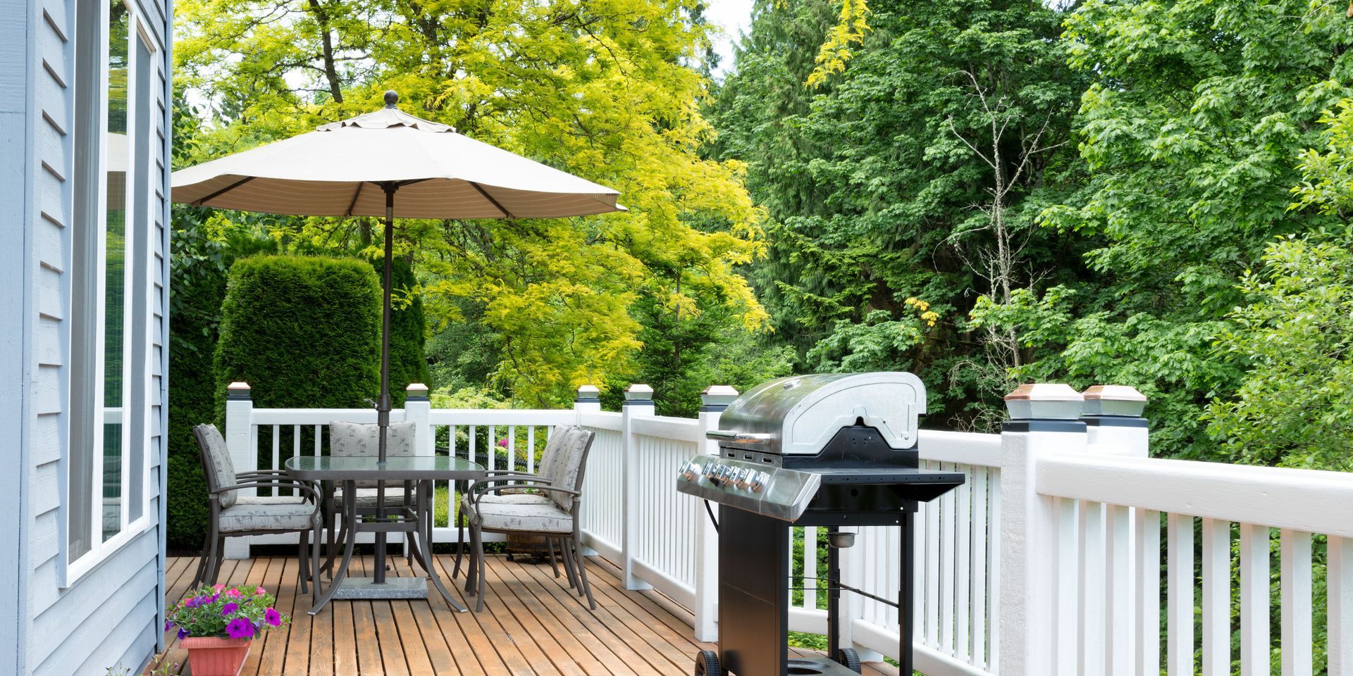 Backyard deck with a grill, table, and umbrella. Lush green trees in the background.