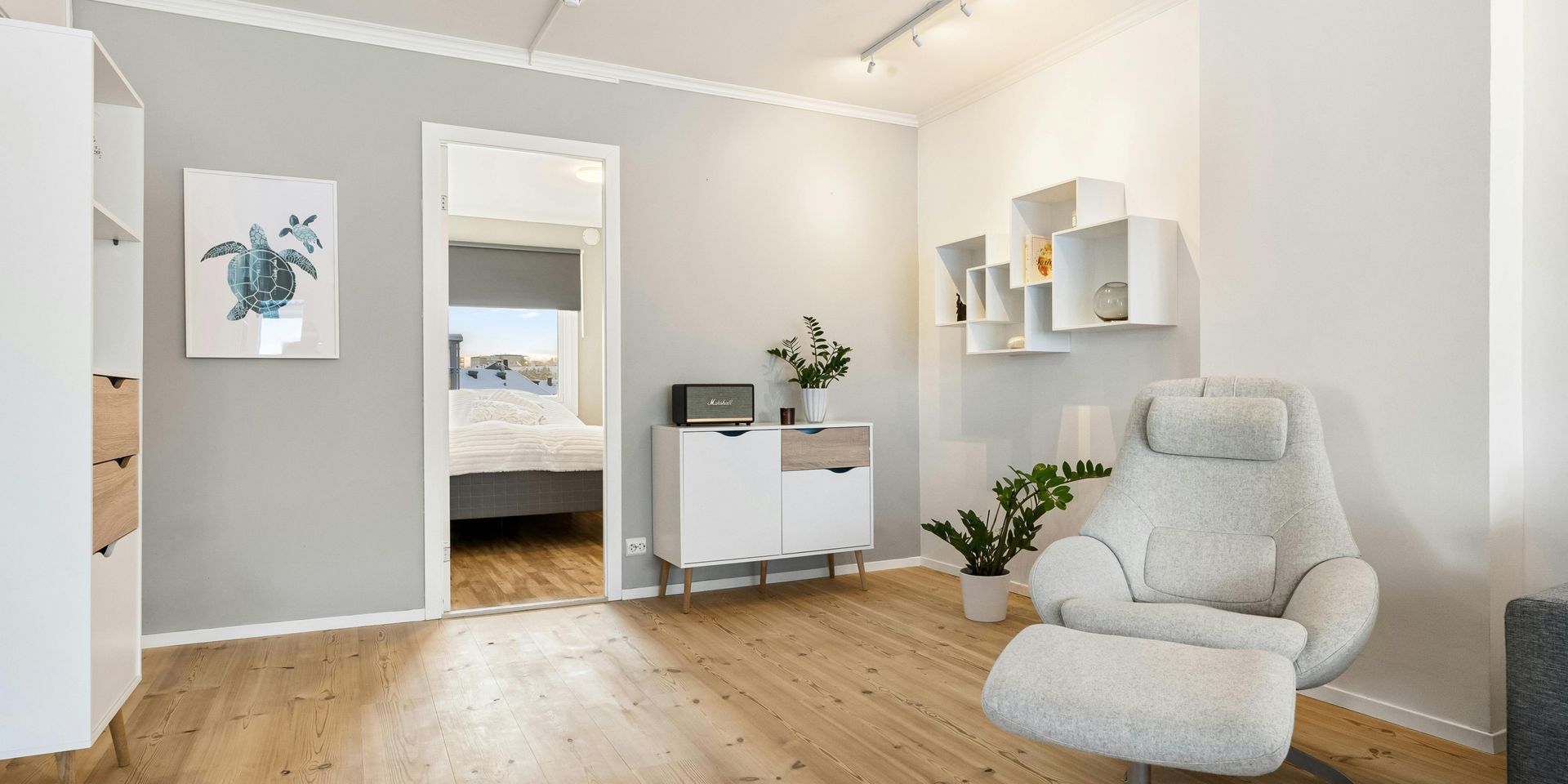 A light-filled living room with a recliner, artwork, and an open doorway to a bedroom.