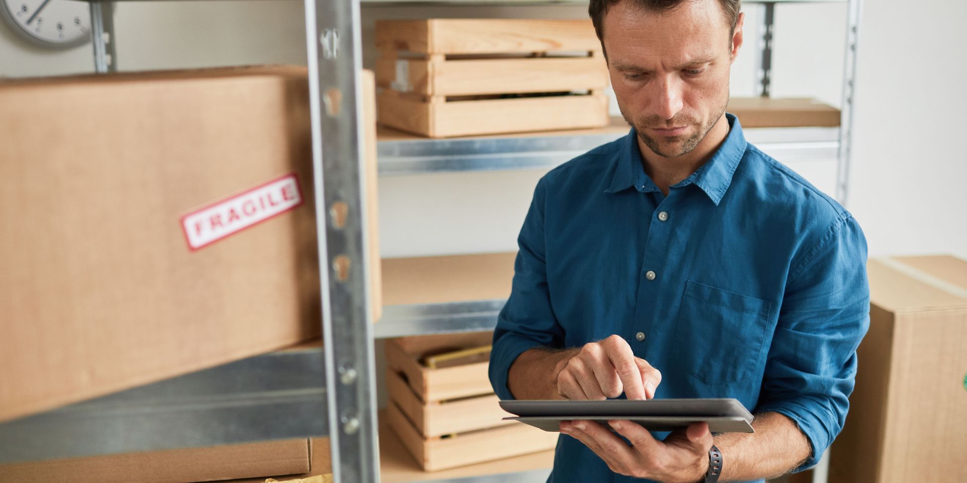 Man in blue shirt using a tablet in a warehouse; shelves with boxes in background.