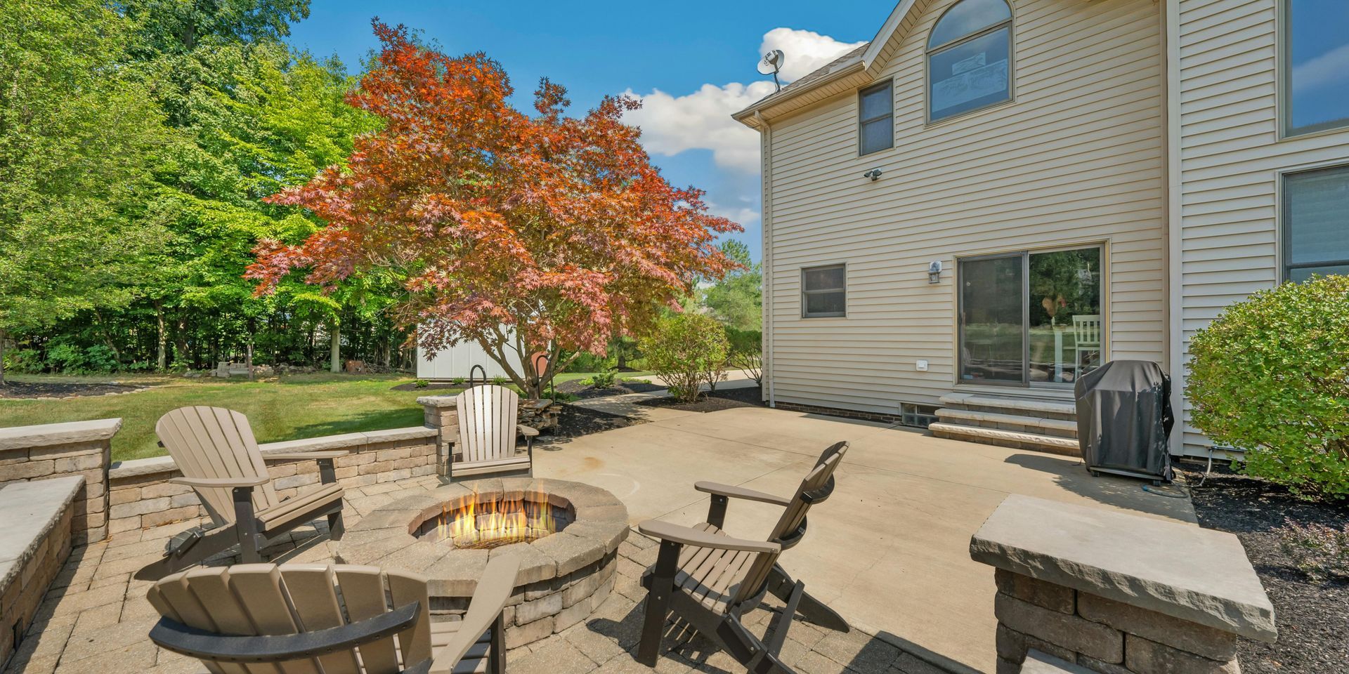 Patio with fire pit, chairs, and house in the background. Bright autumn tree and green grass. Sunny day.