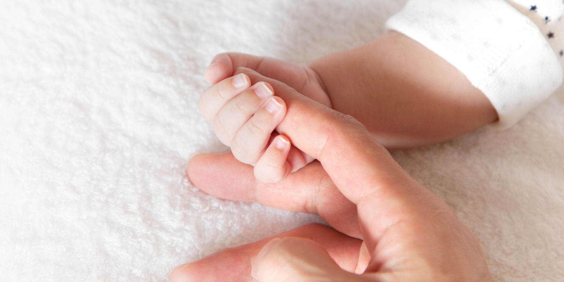 A small baby hand grasping an adult's finger, resting on a white, soft surface.