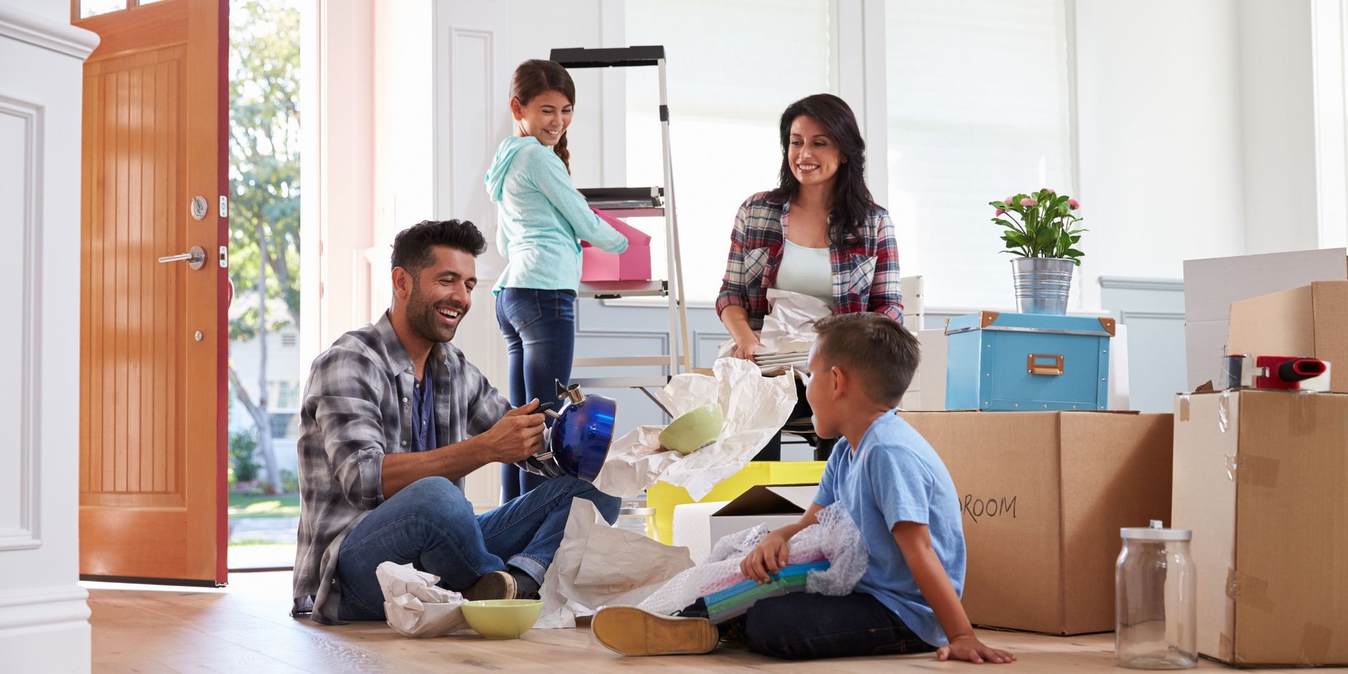 Family unpacking boxes in a new home. Dad sits on floor, mom standing, children helping.