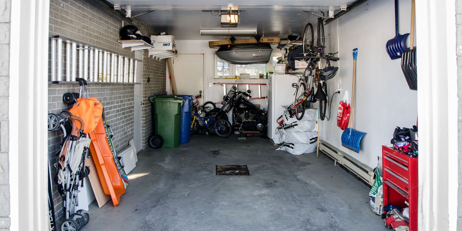View into a cluttered garage. Snowboards, bikes, and tools are stored inside, along with trash cans and other items.
