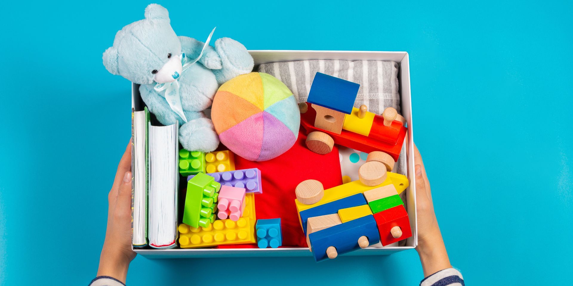 A person is holding a box filled with toys on a blue background.