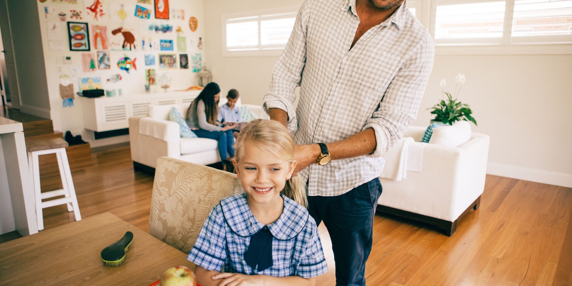 Father braiding his daughter's hair in their bright living room. Family sitting on a couch in the background.