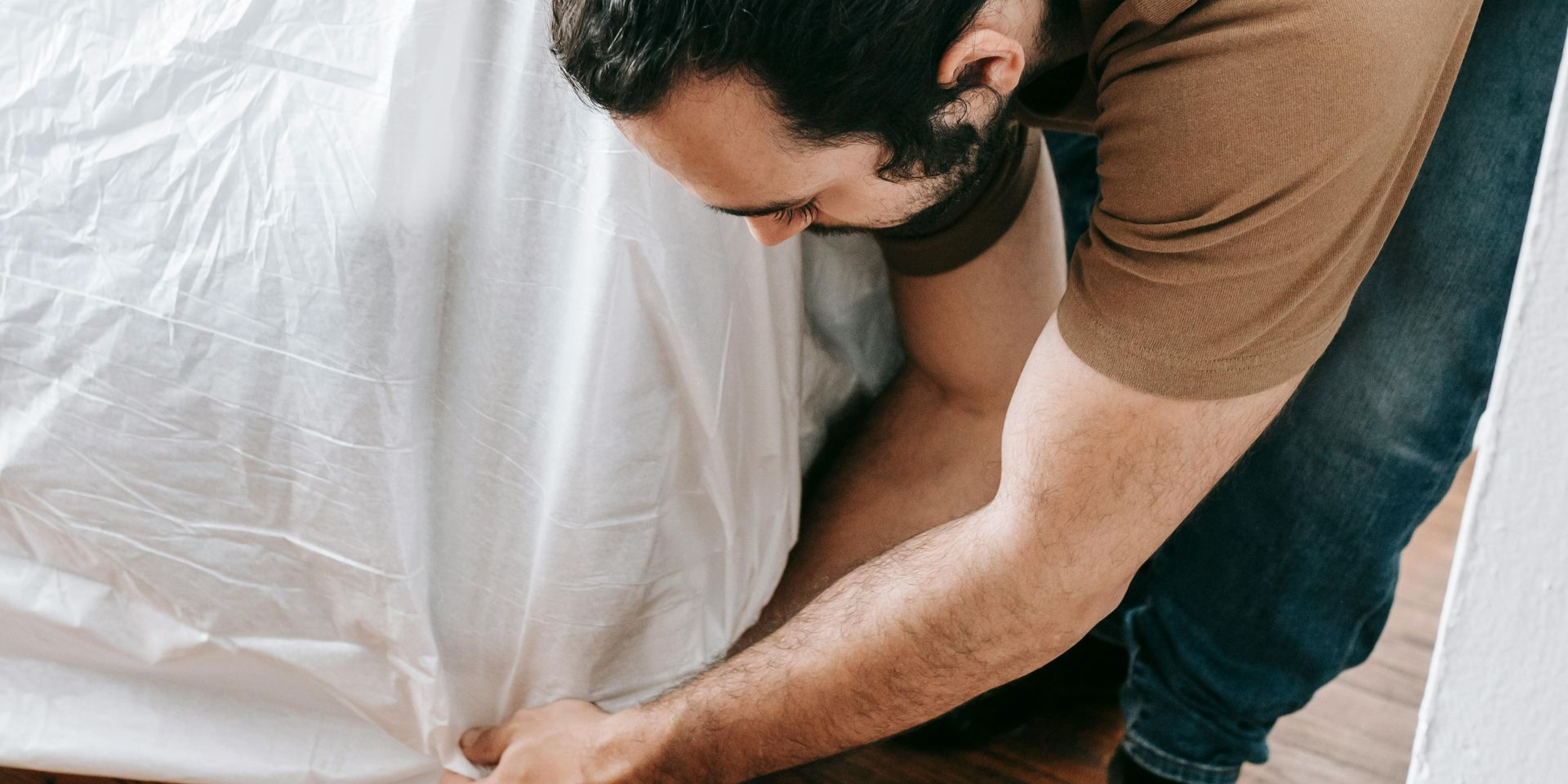 Person tucking a white sheet onto a bed in a room with wood flooring.