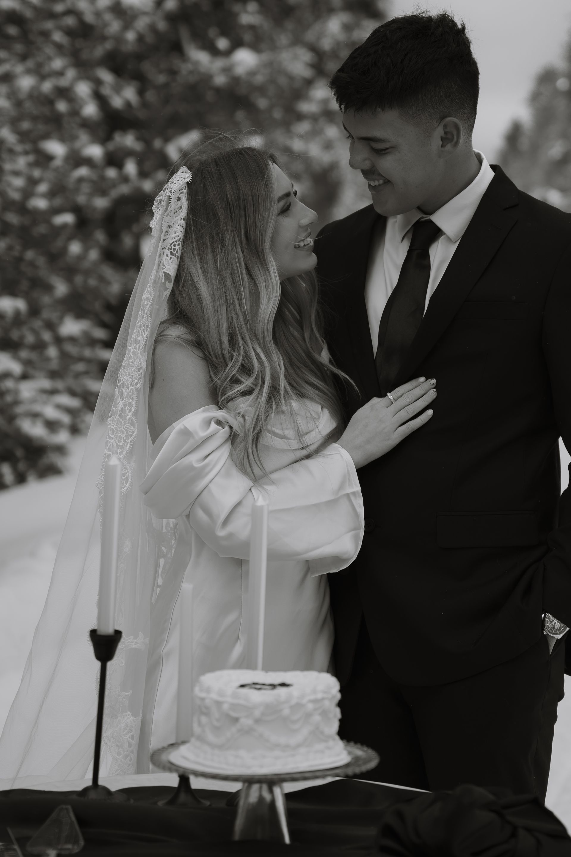 A bride and groom are standing next to a cake in the snow.