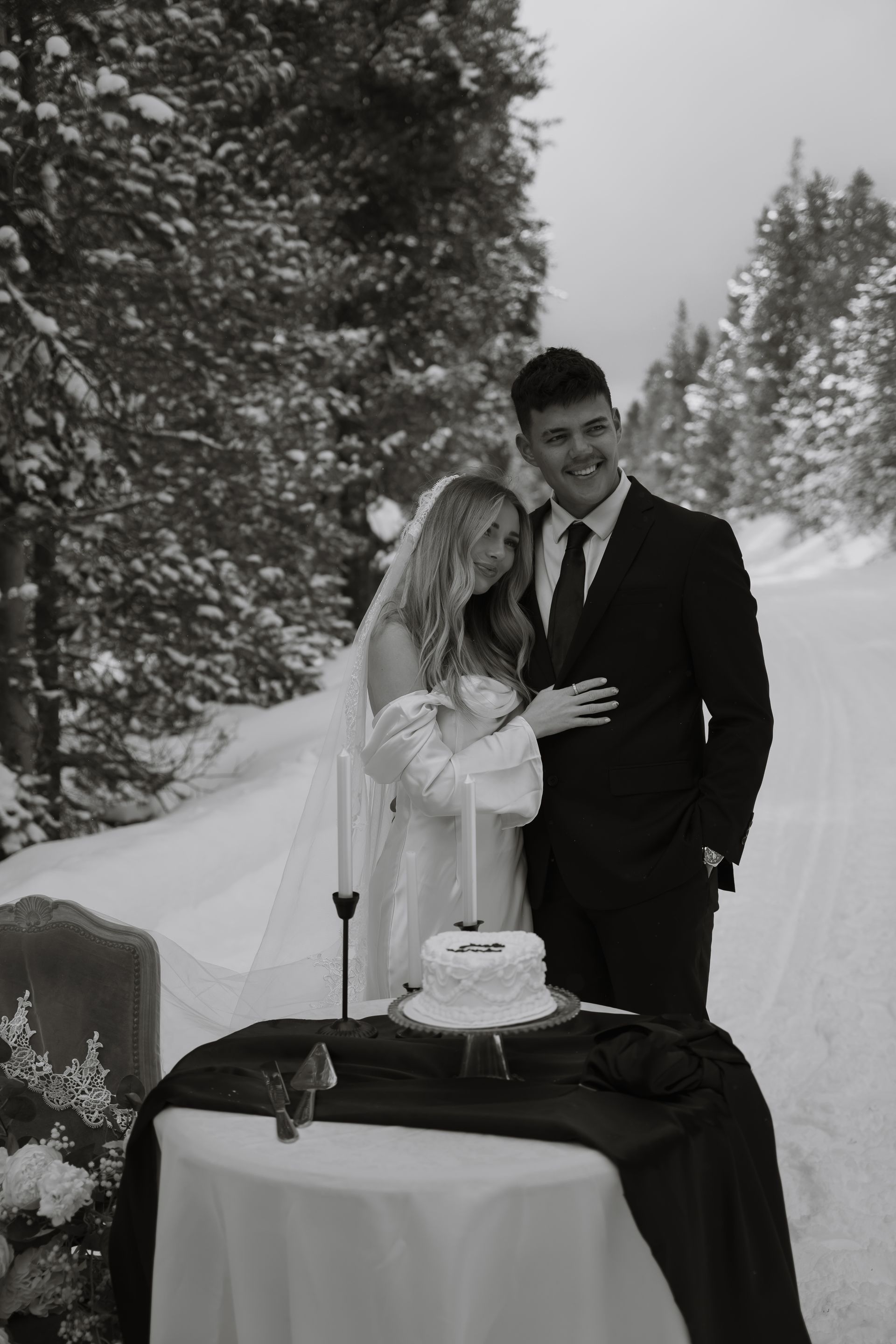 A bride and groom are standing next to a cake on a table in the snow.