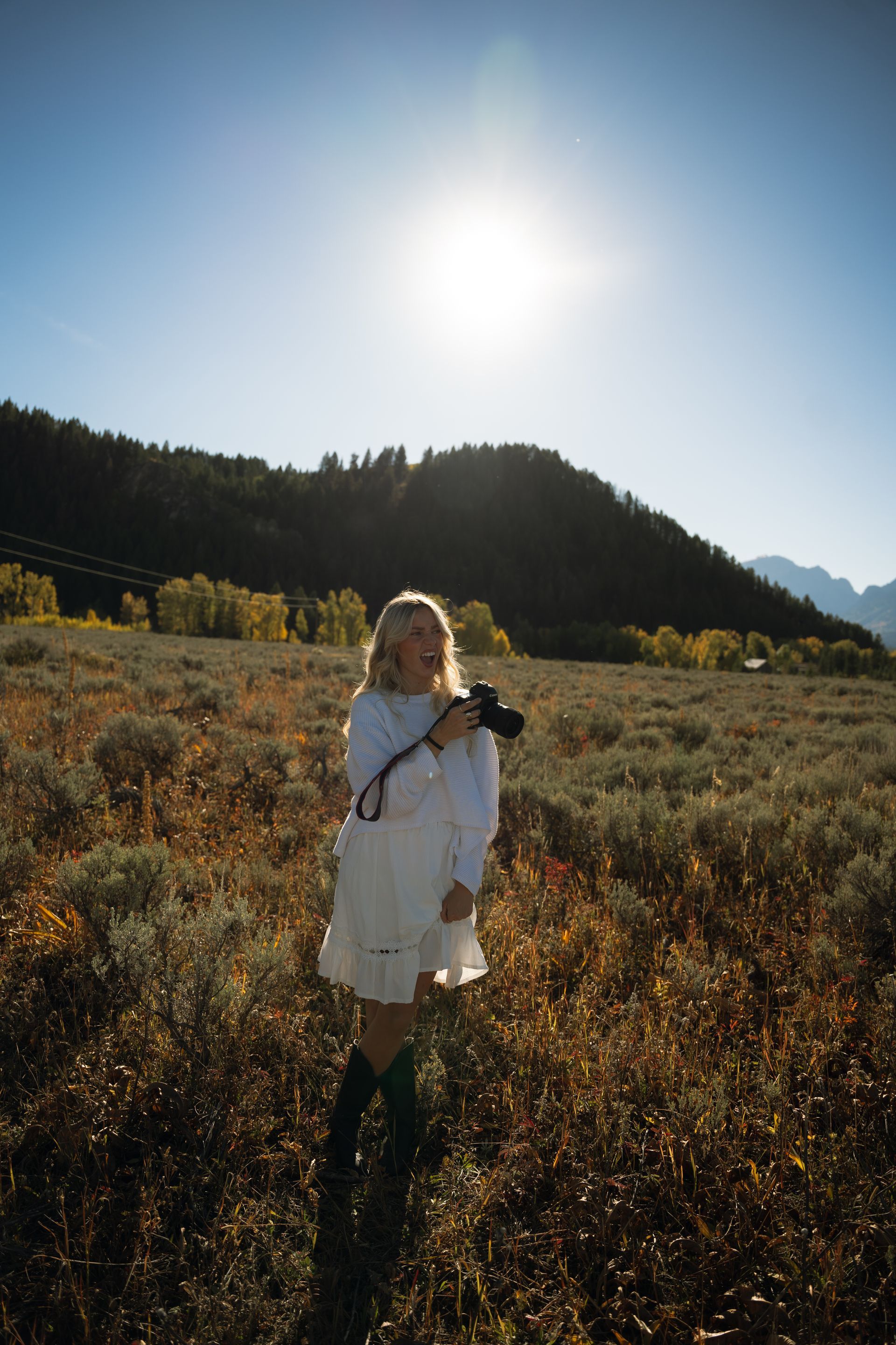 A woman in a white dress is standing in a field holding a camera.