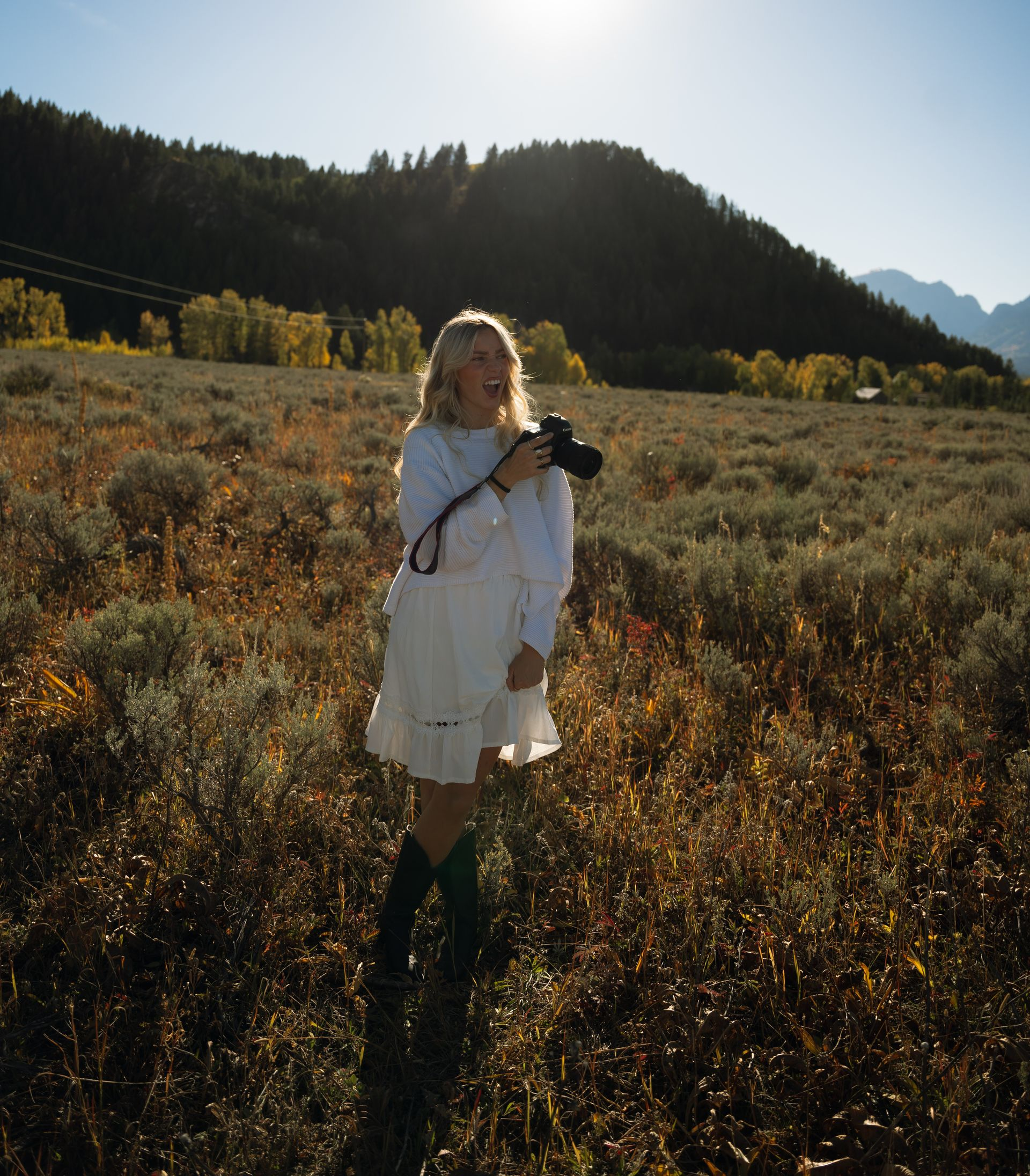 A woman in a white dress is standing in a field holding a camera.