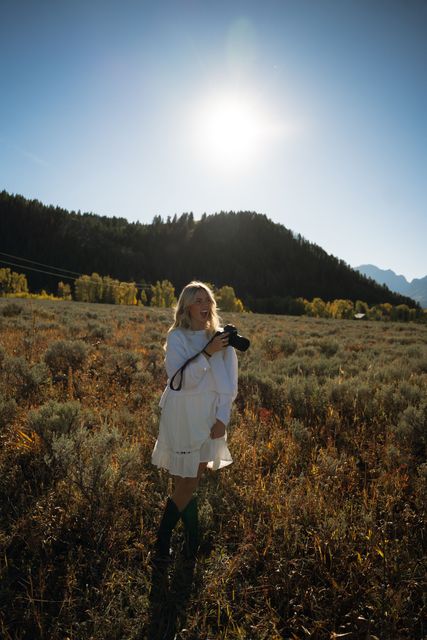 A woman in a white dress is standing in a field holding a camera.
