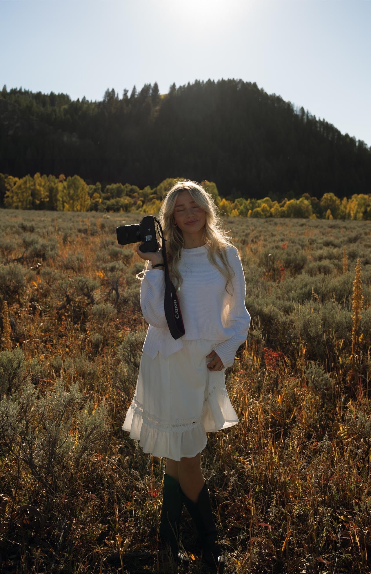 Blond woman in white dress and boots holds camera in field with mountain backdrop. Sunlit scene.