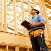 Construction worker in a white hard hat inspecting wooden framing, holding a clipboard, looking up. Wearing tool belt, outdoors.