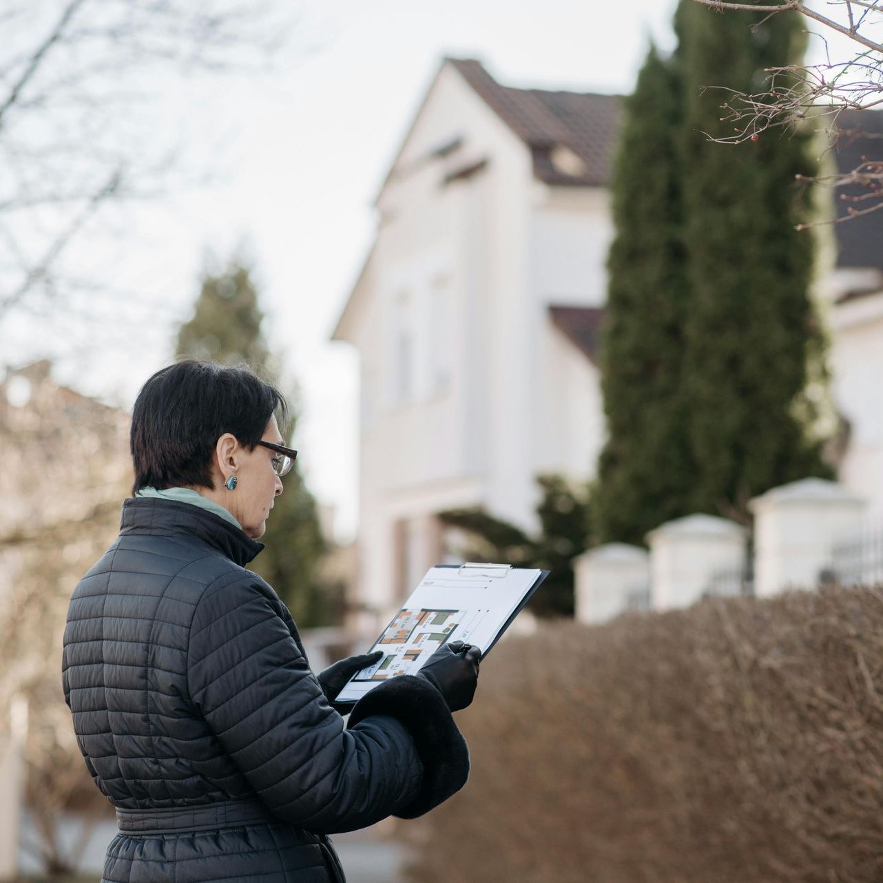 Woman in black coat and gloves holding a clipboard with photos, outside a white house.