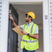 A construction worker wearing a hard hat and vest inspects a doorway, holding a clipboard, with the number 11 visible.
