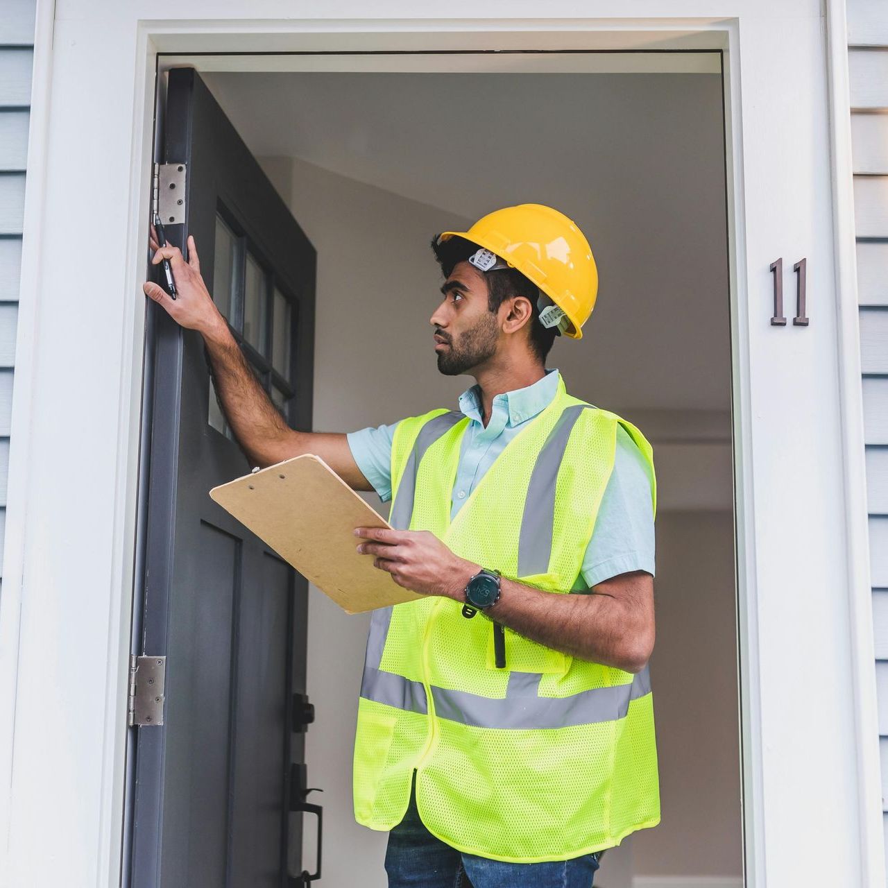 A construction worker wearing a hard hat and vest inspects a doorway, holding a clipboard, with the number 11 visible.