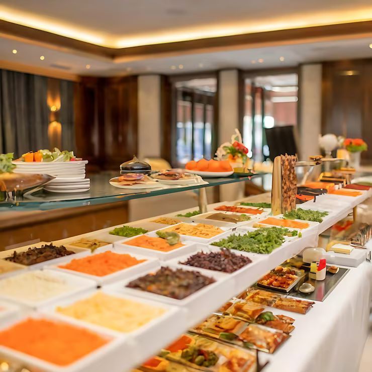 Buffet with a variety of foods in white trays, indoors, with a warm-toned setting.
