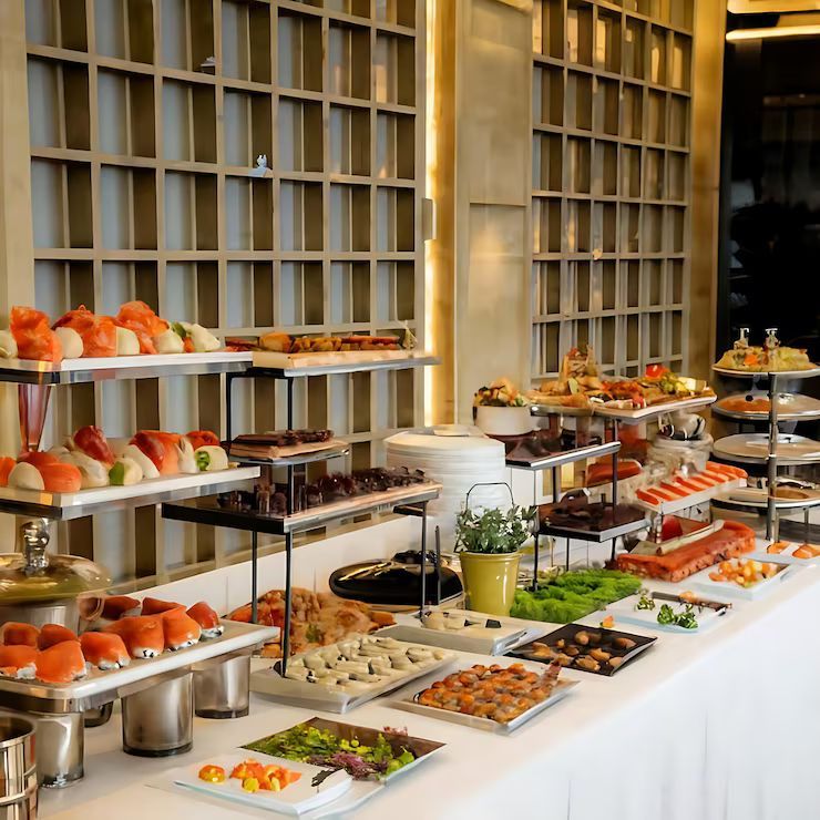Buffet table with various sushi, appetizers, and desserts on tiered platters. White tablecloth, neutral background.