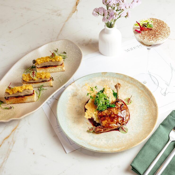 Gourmet meal: duck leg with mashed potatoes, appetizers, tiramisu, and flowers on a marble table.