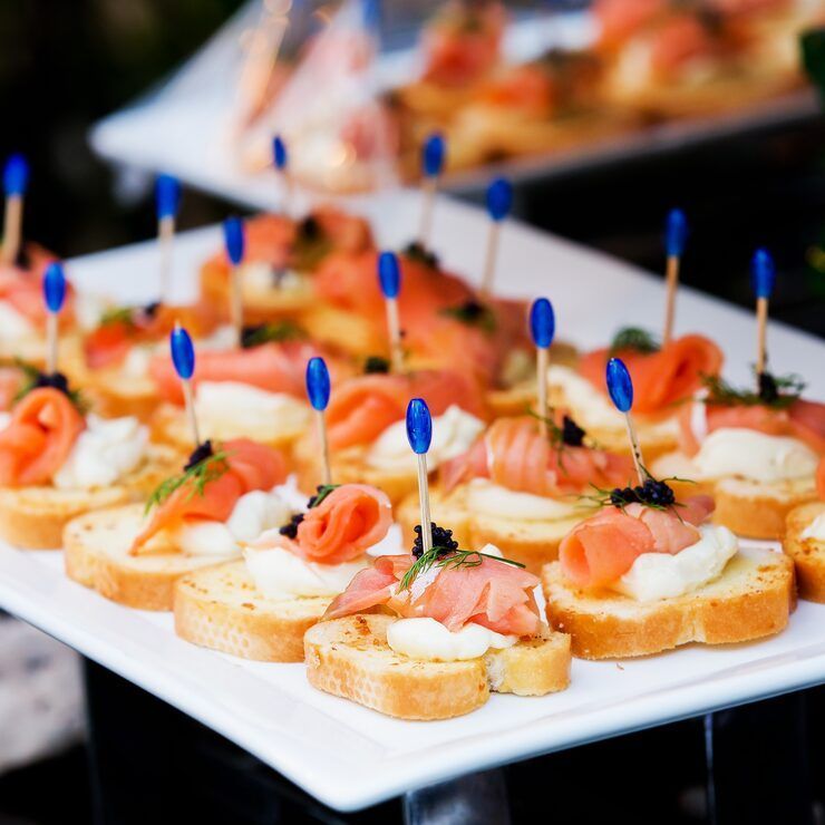 Smoked salmon appetizers on toasted bread, topped with cream cheese, caviar, and dill, secured with blue toothpicks.