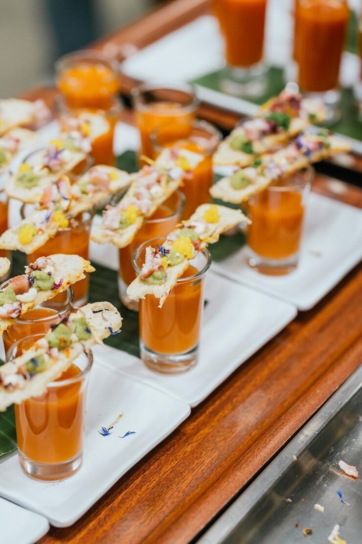 Small glasses of orange soup topped with crackers and garnish, arranged on white rectangular plates on a wooden table.
