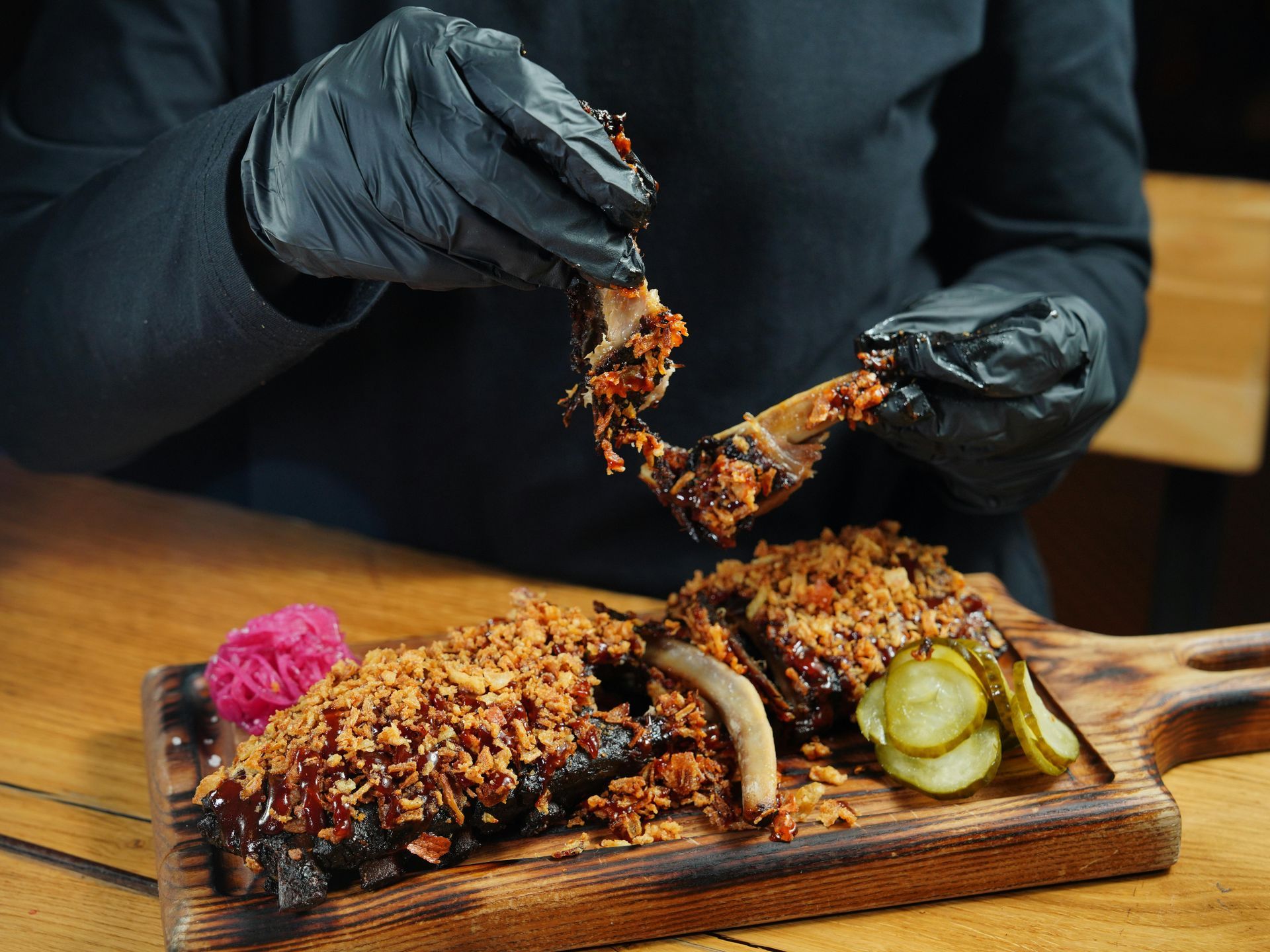 Person in black gloves pulling apart a cooked rib, on a wooden board with sides, pickles, and red cabbage.