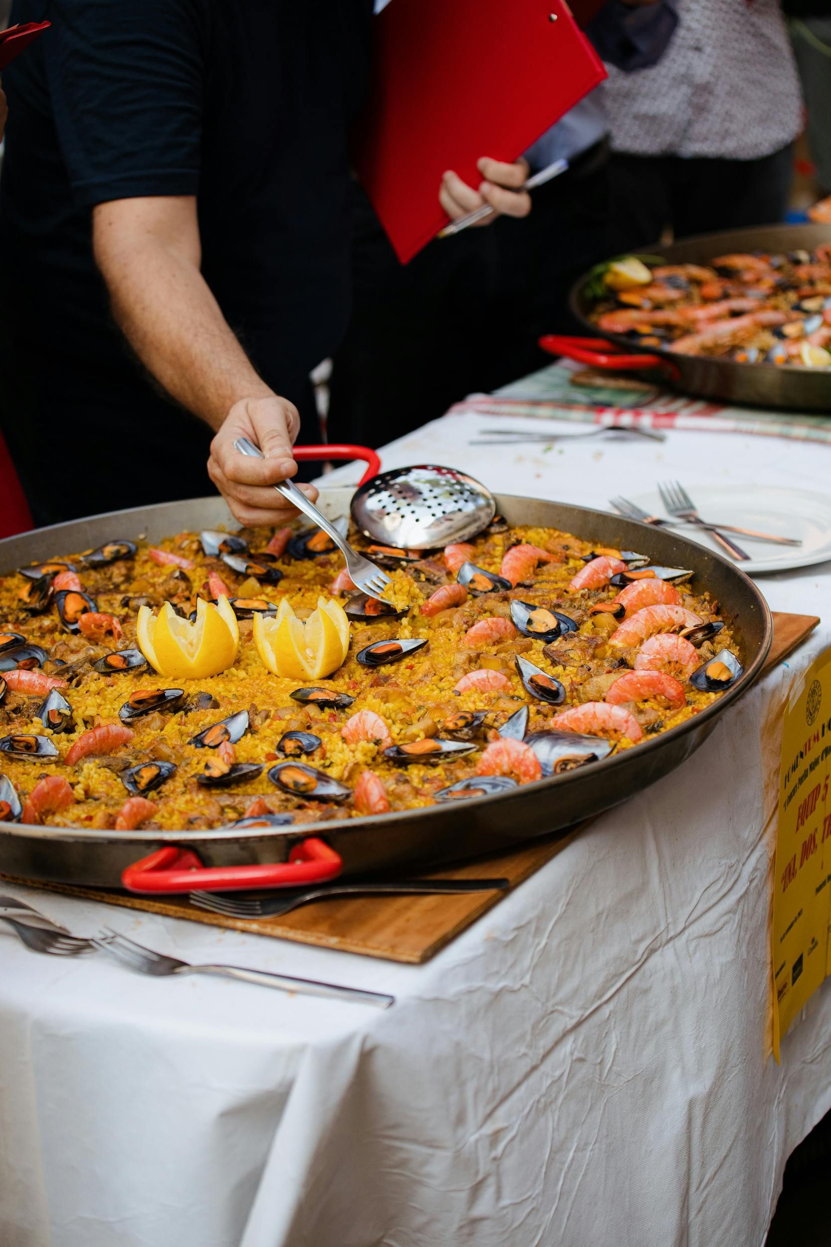 Chef serving paella with seafood, lemon garnish, from a large pan on a white tablecloth.