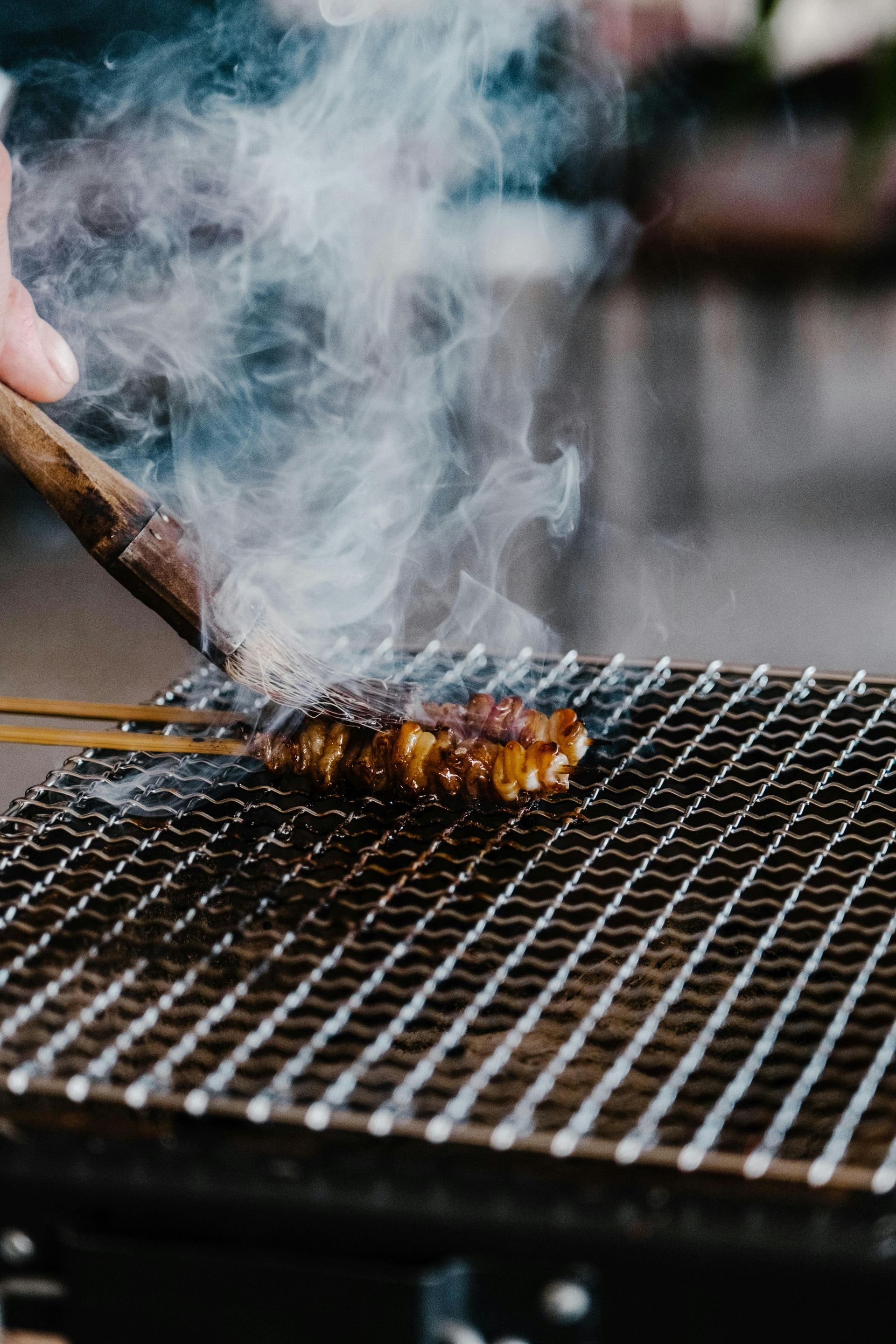 Meat cooking on a grill suspended over a fire; tongs in hand.