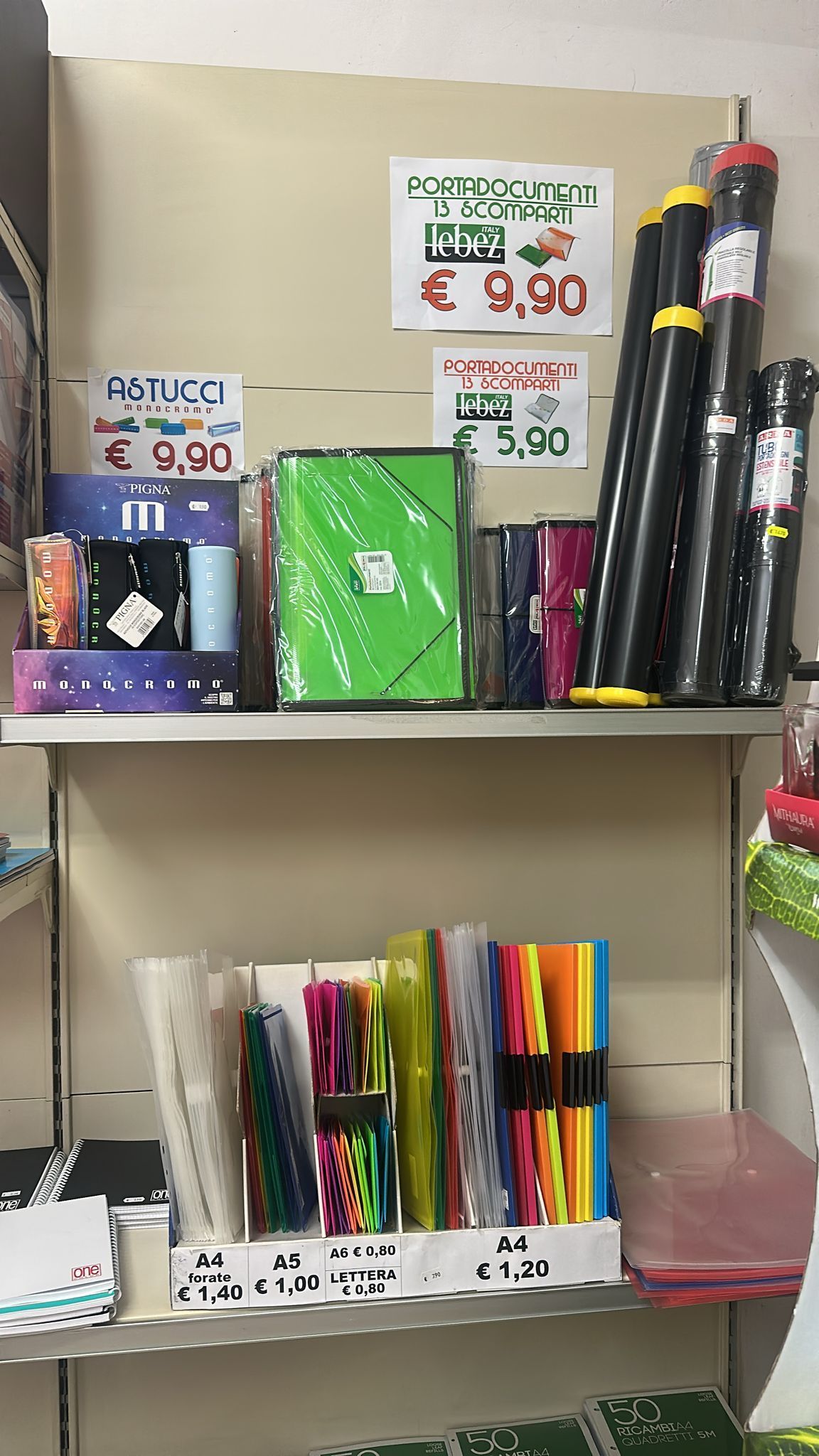 Scaffali con articoli di cancelleria, tra cui quaderni, cartelle e tubi porta-poster. I prezzi sono visibili.