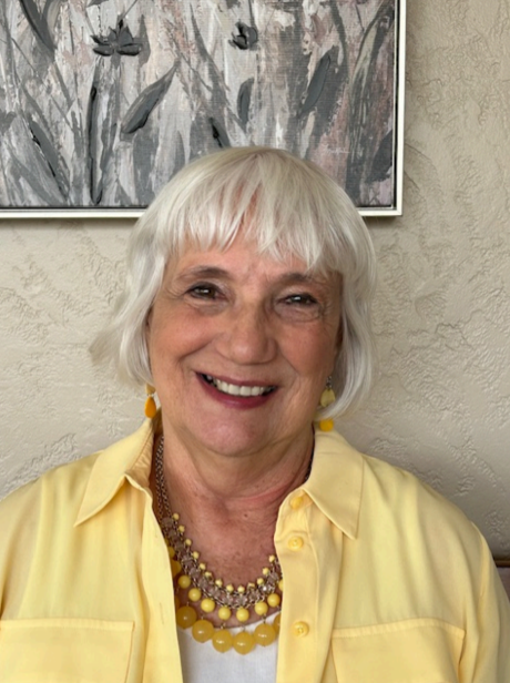 A woman in a yellow shirt is smiling in front of a painting.