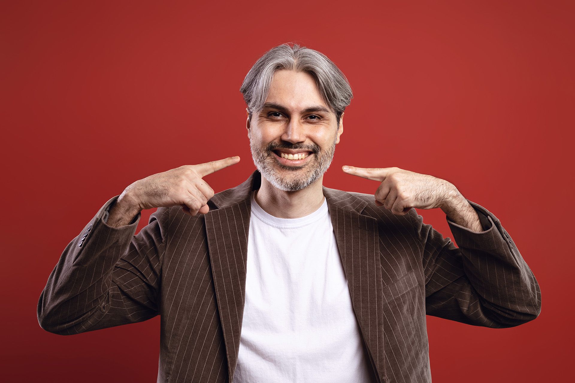 Man with graying hair and beard smiles, pointing at his smile; red backdrop.