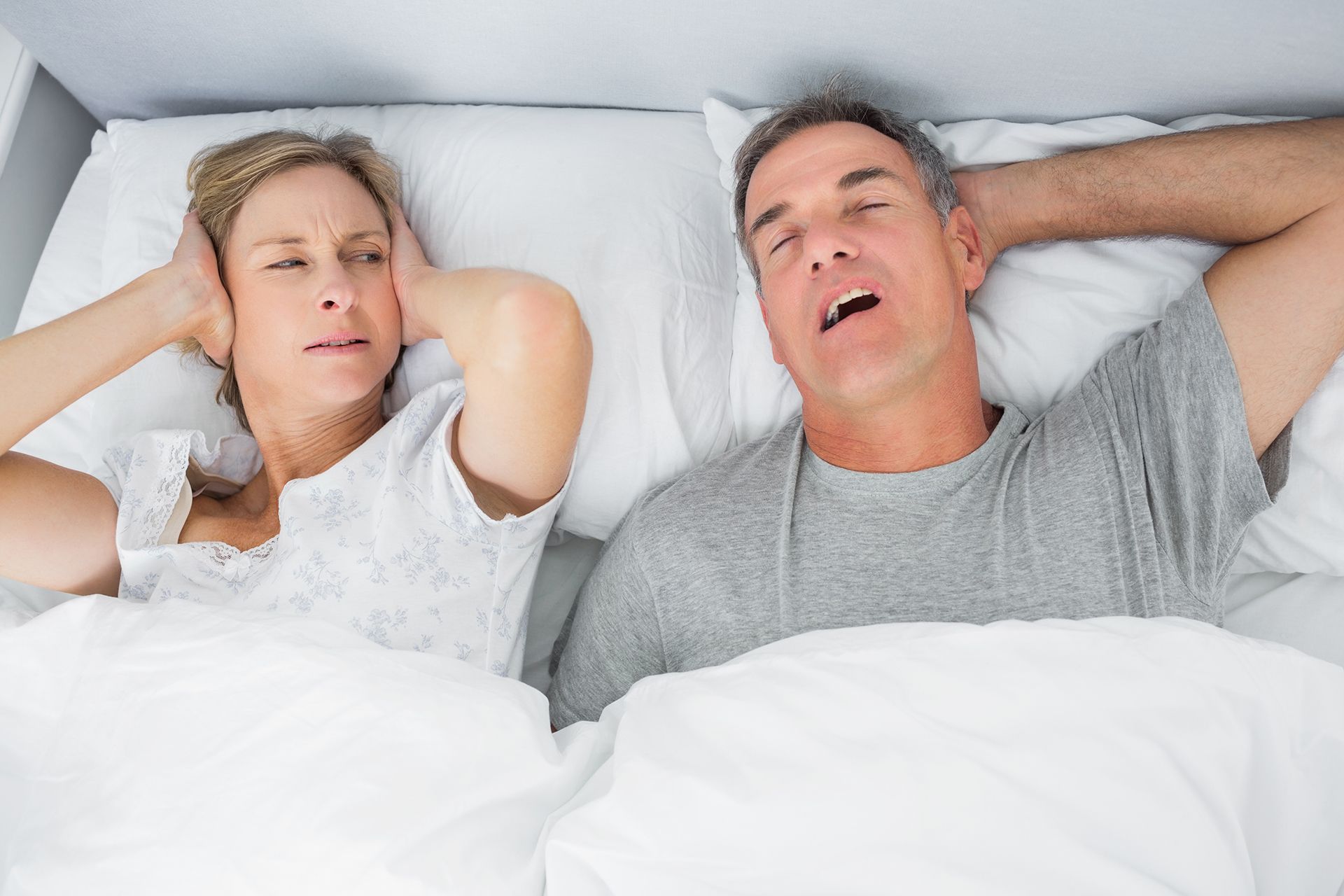 Woman covering ears, man snoring in bed.