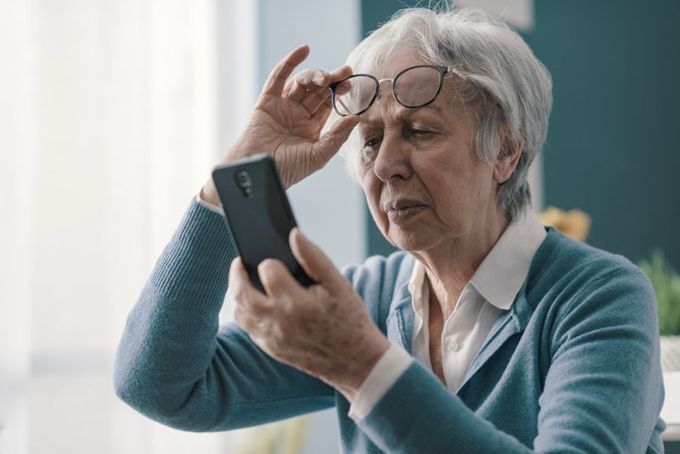 An older woman with gray hair, wearing a blue sweater and white shirt, holds her glasses above her eyes while squinting at a smartphone, appearing to have difficulty seeing the screen.