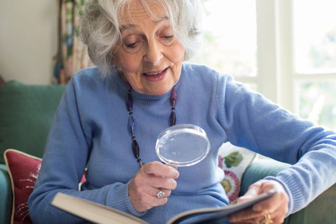 An elderly woman is sitting on a couch reading a book with a magnifying glass.