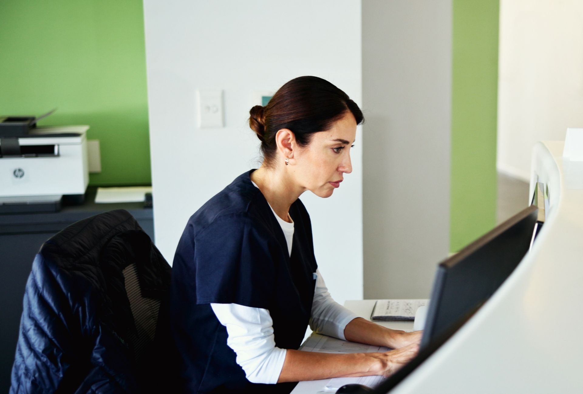 A woman is sitting at a desk in front of a computer scheduling a patient appointment.
