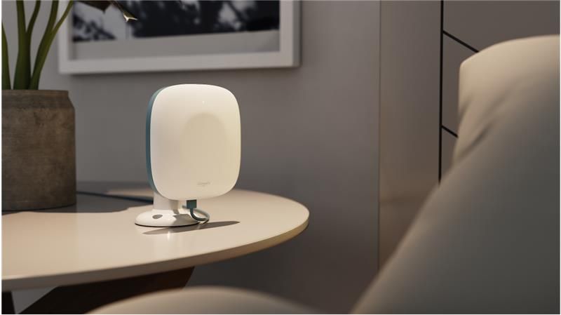 A tabletop remote patient monitoring device sits on a round surface next to a cushioned chair. The device has a sleek, white, rounded design with a soft blue edge and is connected via a cable to its base. Designed for contactless collection of patient vital signs, it blends discreetly into the modern, softly lit room, suggesting a comfortable healthcare or assisted living environment.