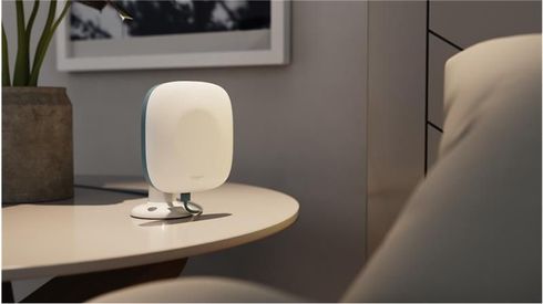 A tabletop remote patient monitoring device sits on a round surface next to a cushioned chair. The device has a sleek, white, rounded design with a soft blue edge and is connected via a cable to its base. Designed for contactless collection of patient vital signs, it blends discreetly into the modern, softly lit room, suggesting a comfortable healthcare or assisted living environment.