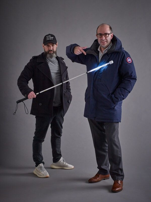 Two men standing together in a studio setting. One man with a beard, wearing a black coat, gray shirt, cap, and sneakers, holds a white mobility cane. The other man, wearing glasses, a navy winter jacket, dress pants, and brown shoes, points to the illuminated cane while smiling at the camera.
