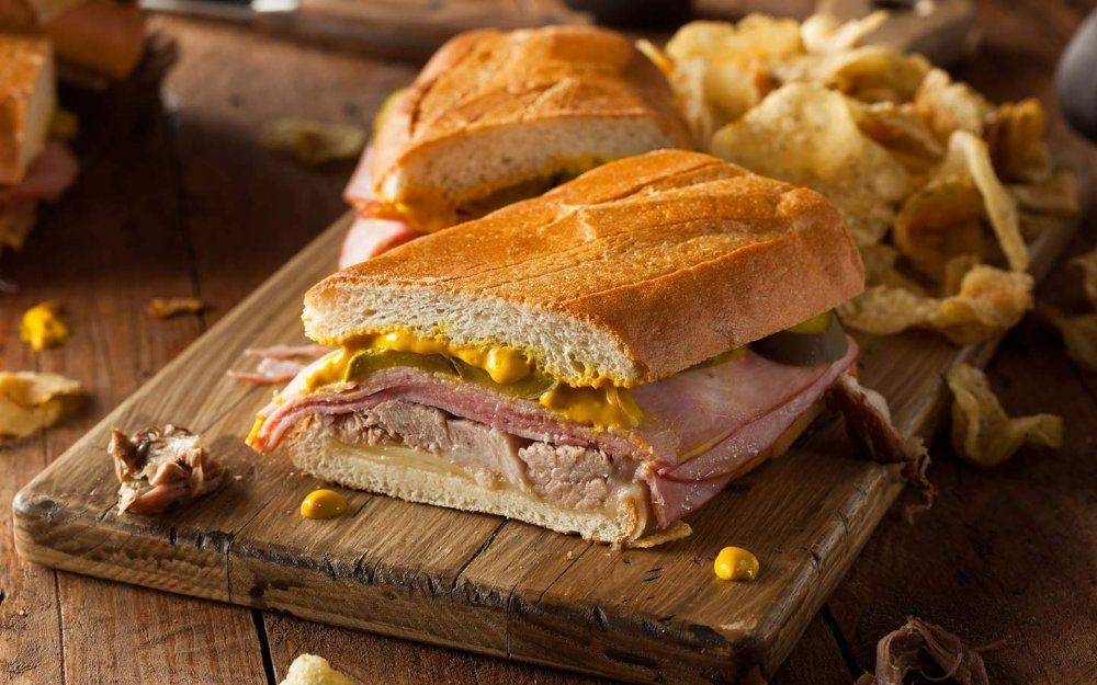 A cuban sandwich is sitting on a wooden cutting board next to potato chips.