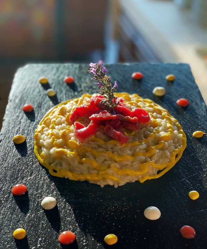 Risotto con salsa gialla, guarnito con carne rossa e fiori su una lavagna nera, decorato con gocce colorate.