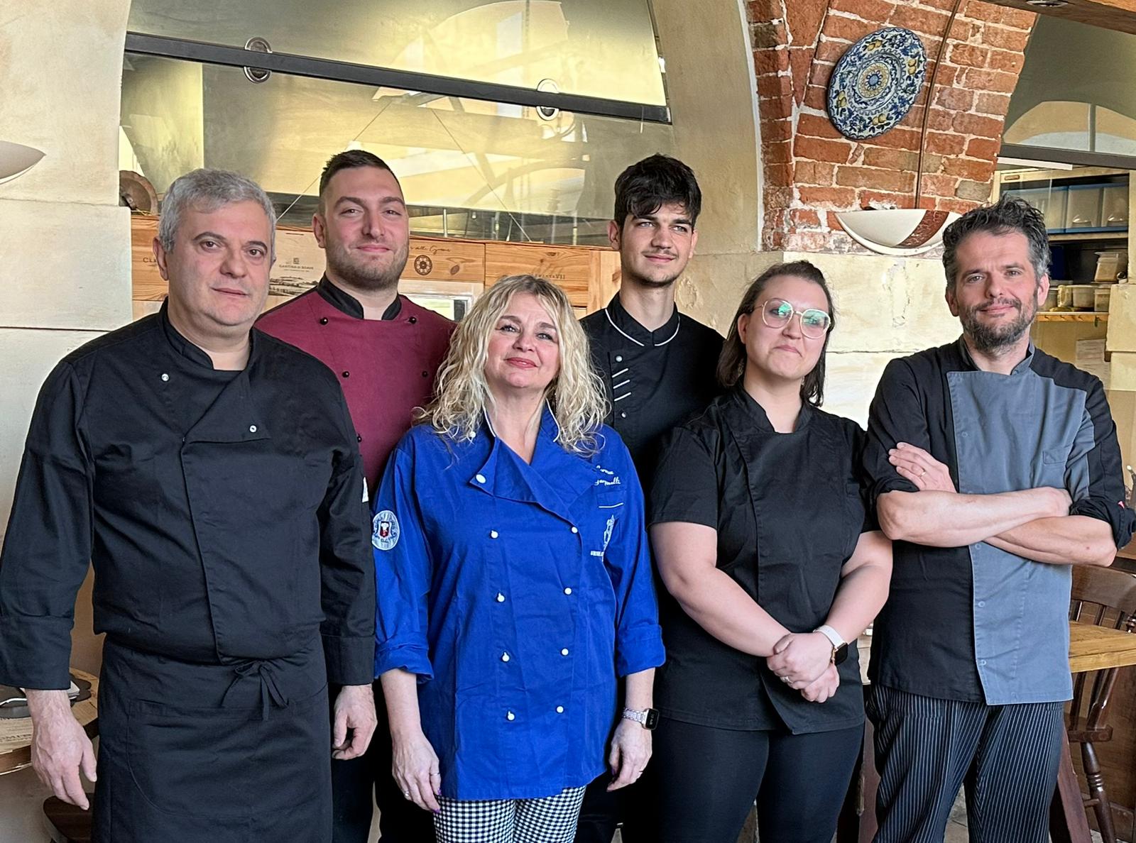 Gruppo di sei membri dello staff di un ristorante in tenuta da chef, in posa all'interno. Sullo sfondo un muro di mattoni.