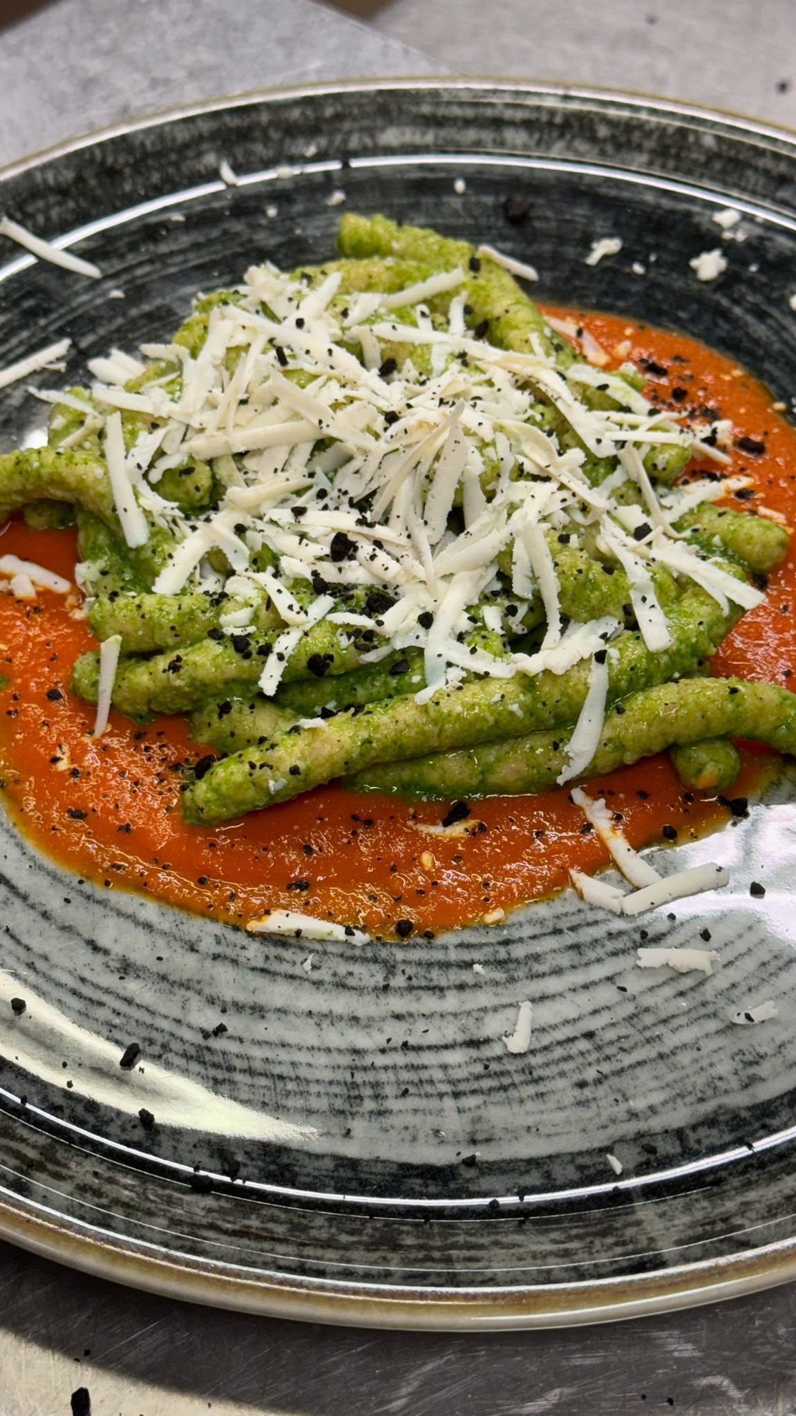 Pasta verde con formaggio grattugiato su salsa rossa, servita su un piatto con bordo grigio.